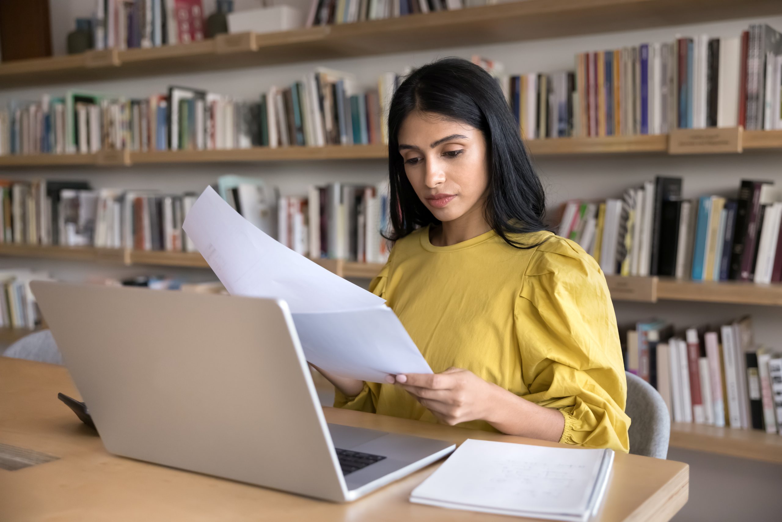Woman in yellow blouse reviewing documents with concerned expression while working on laptop in library setting, reviewing data to identify instructional gaps.