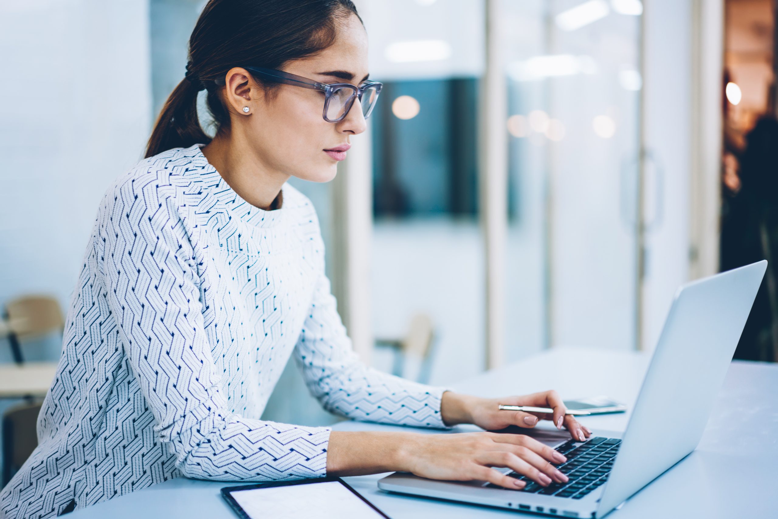 Professional woman with glasses in patterned blouse intently analyzing data on laptop computer in modern office setting, reviewing educational performance metrics.