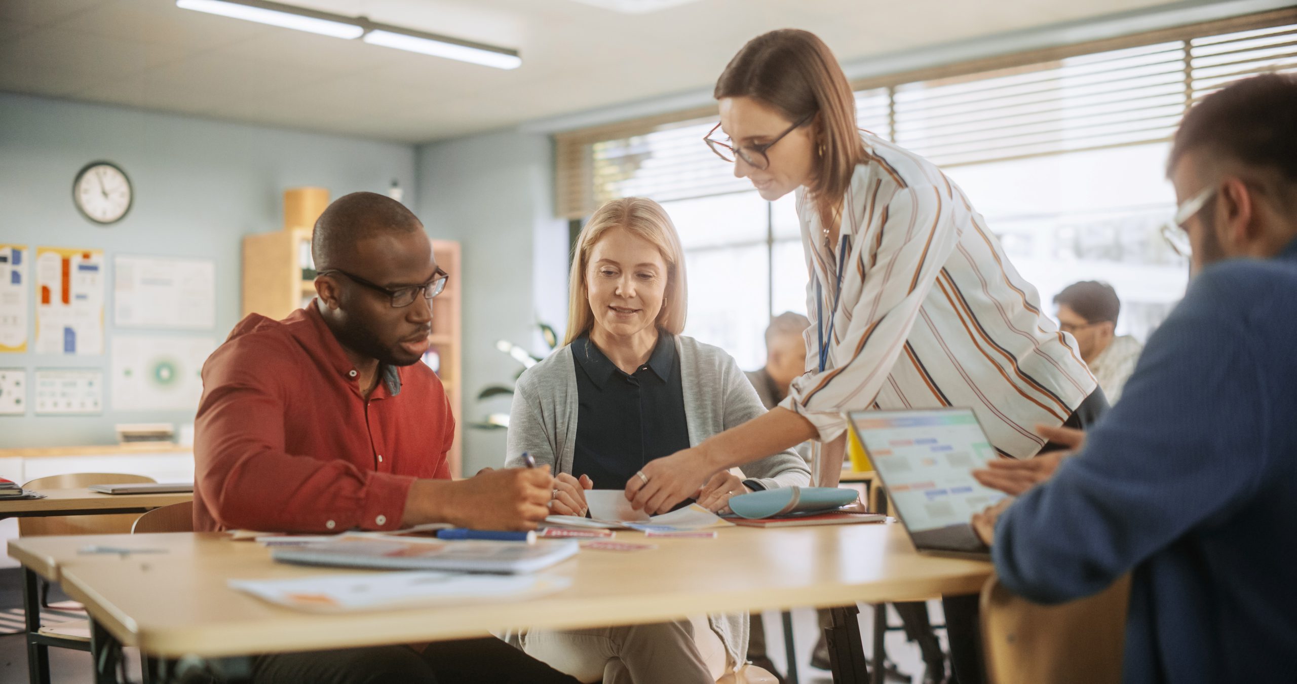 Diverse group of teachers collaborating around table in classroom, with woman in striped blazer pointing to data while colleagues review documents and charts on wall visible in background.