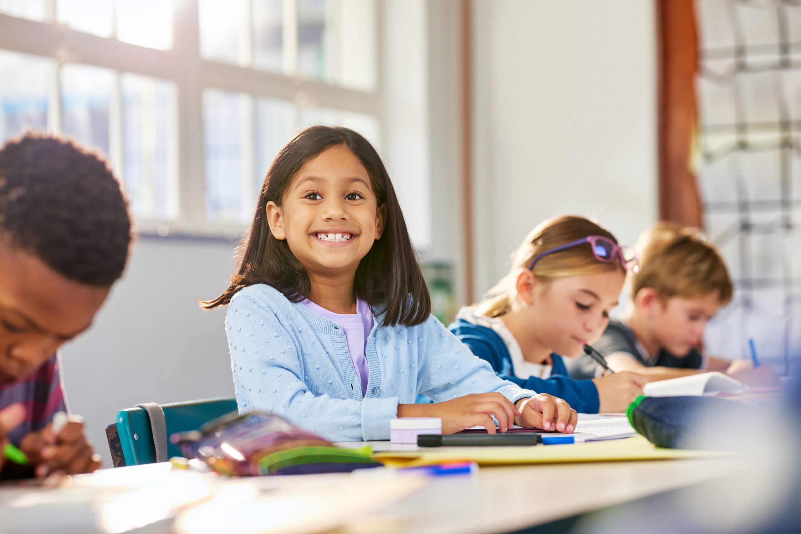 A young girl smiles confidently at her desk during a classroom activity, surrounded by peers working independently.