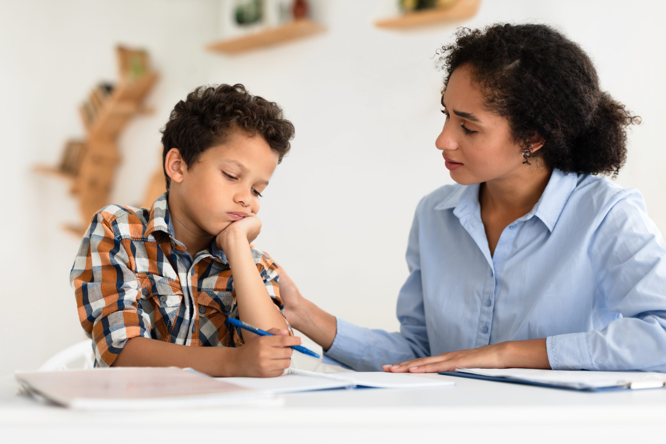 Female teacher with curly hair in blue shirt providing one-on-one support to young boy in plaid shirt who appears discouraged while working on writing assignment at white desk.