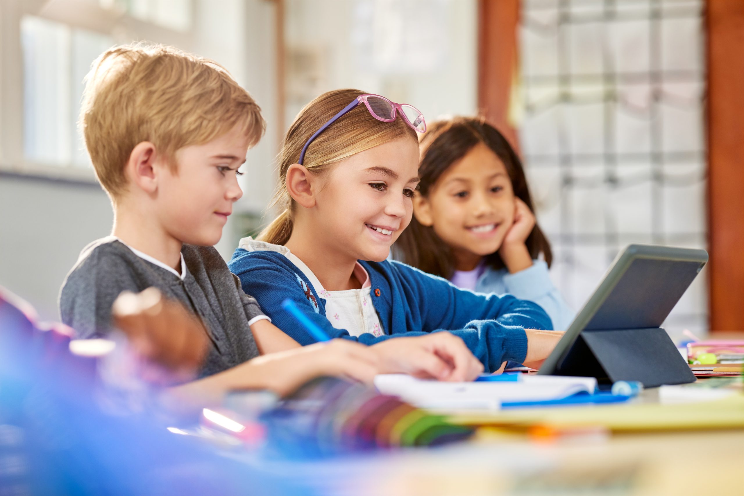 Three primary school students smile as they work together on a tablet in a classroom setting.