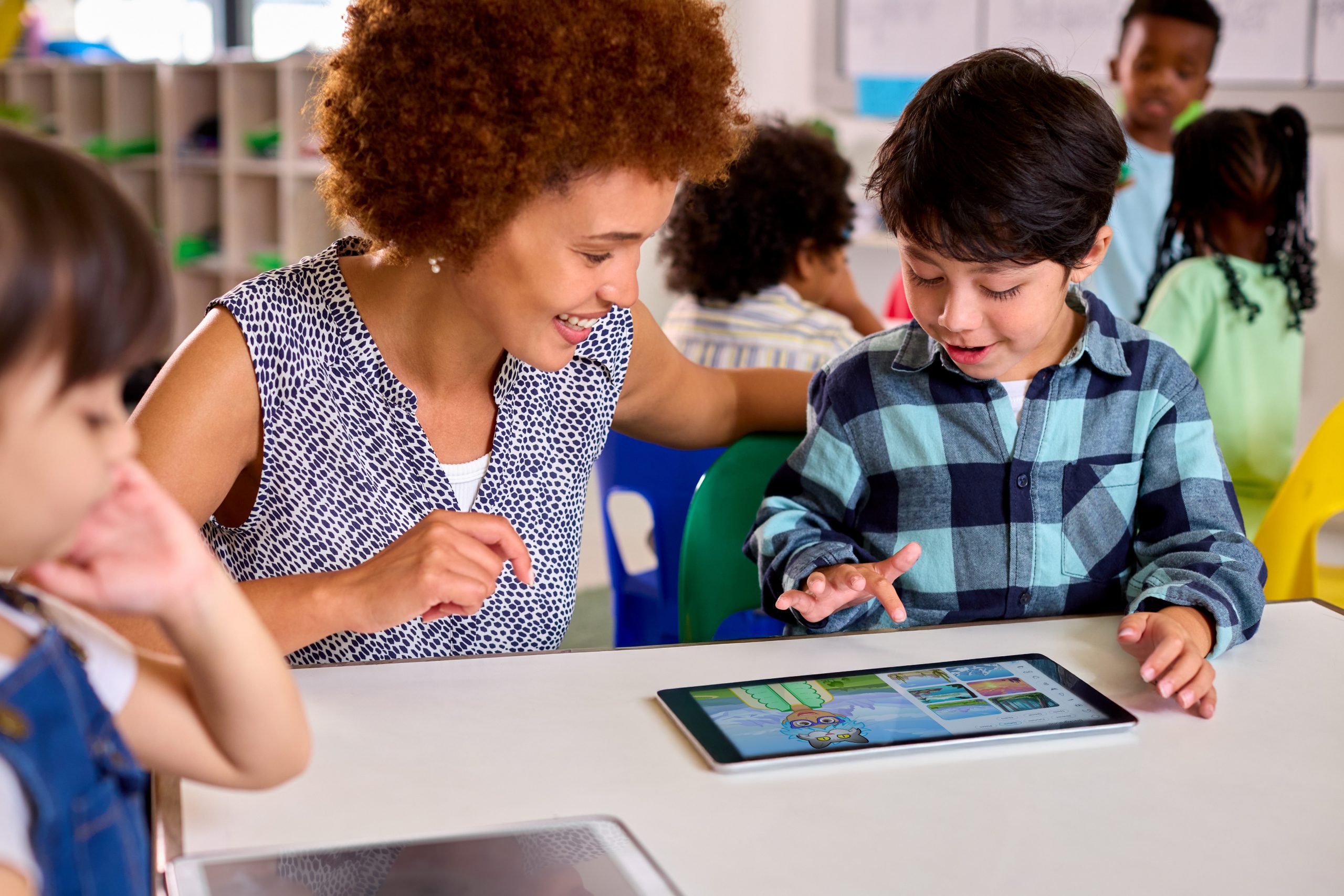 A teacher sits alongside a young student as they explore a learning program together on a tablet, with other students working independently in the background.